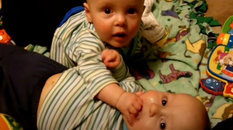 Twin infant brothers are playing together, with one of them sucking the other’s hand like a pacifier