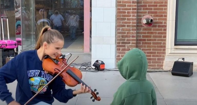 A young child is drawn towards a violinist performing on the street and plants a kiss on her while unable to resist the urge